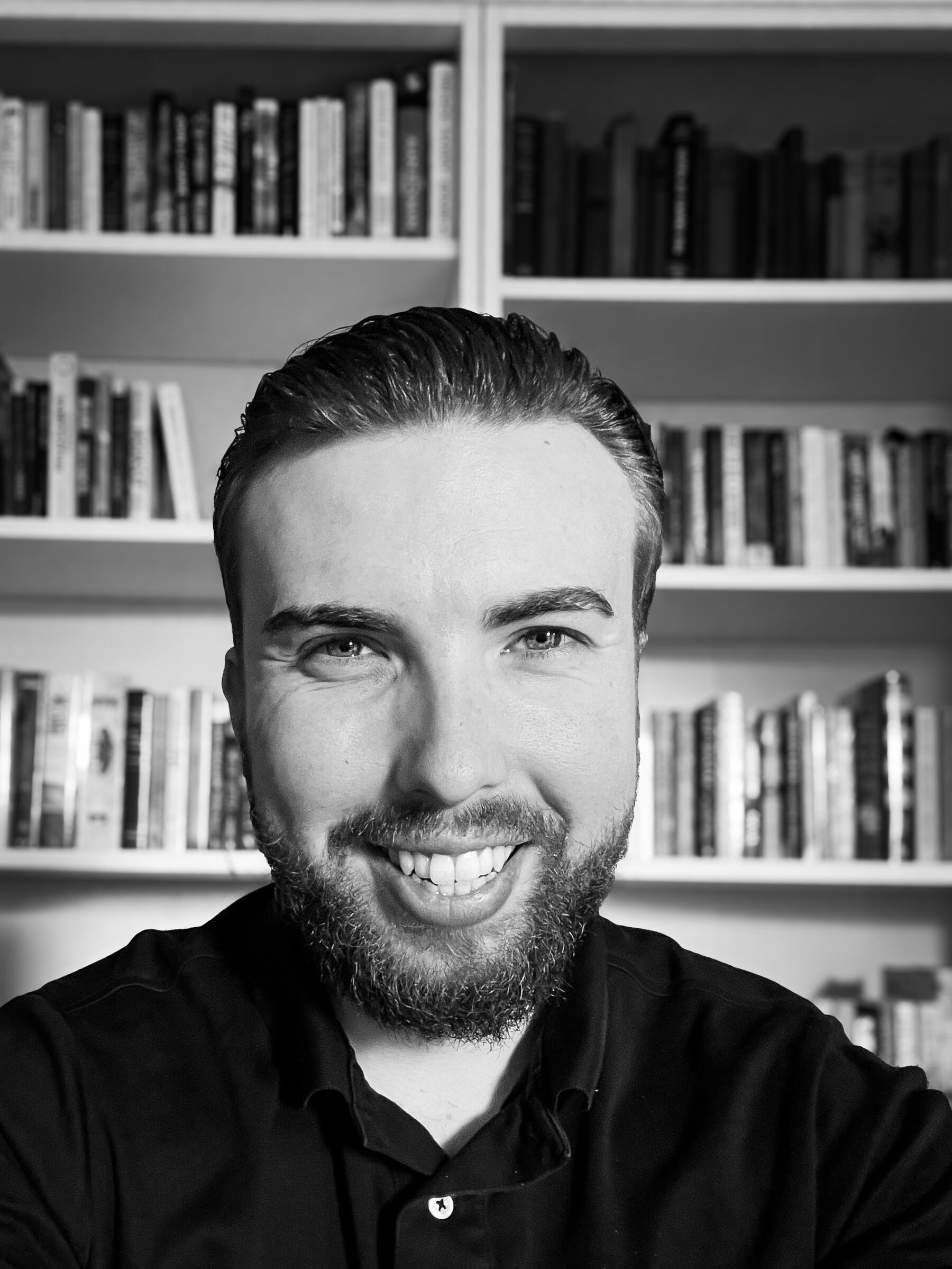 Black-and-white portrait of a smiling man with neatly styled hair and a short beard, wearing a dark collared shirt, sitting in front of a blurred bookshelf background.”